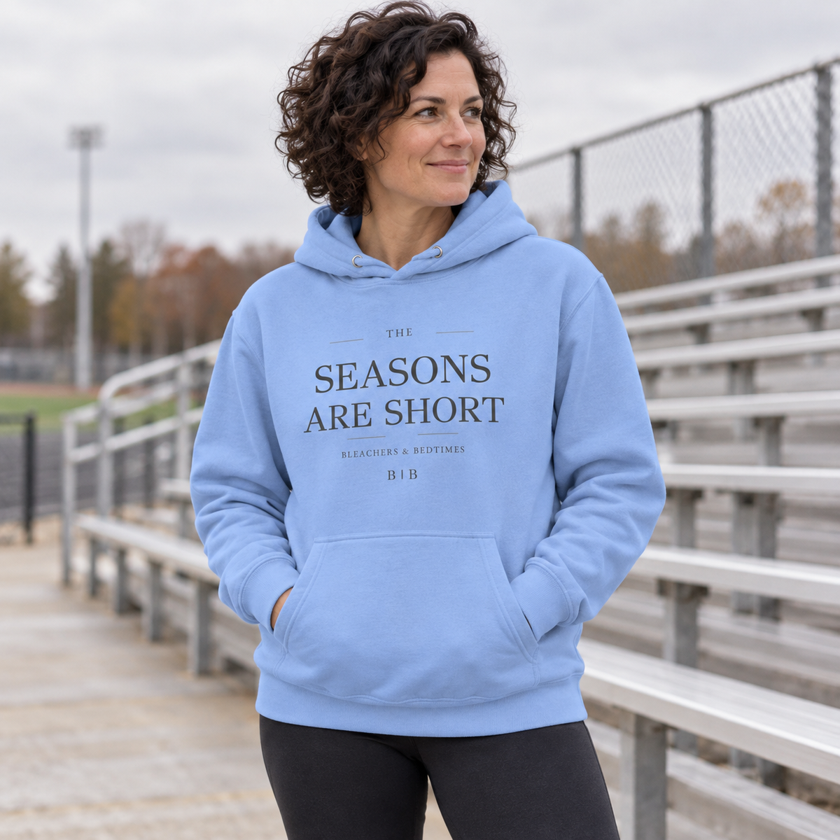 Woman wearing a Carolina blue “The Seasons Are Short” hoodie beside outdoor sports bleachers