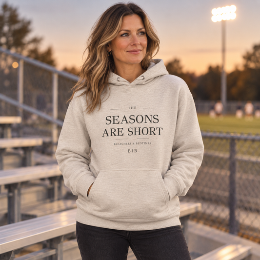 Woman wearing The Seasons Are Short oatmeal hoodie beside outdoor sports bleachers at sunset