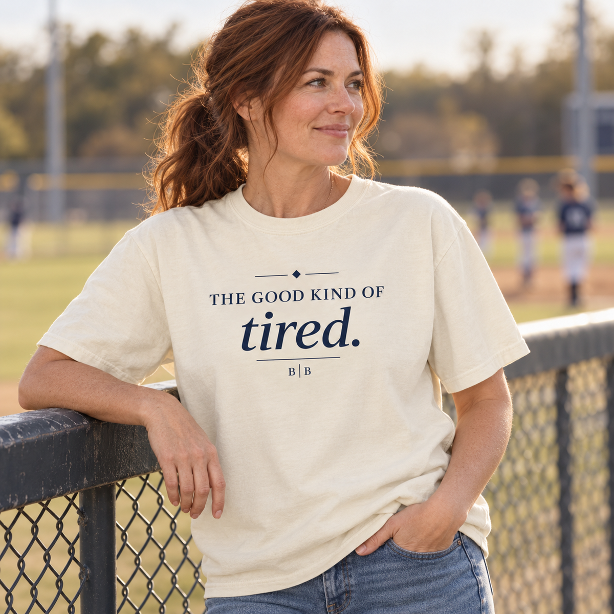 Woman wearing an ivory “The Good Kind of Tired” T-shirt beside a baseball field.