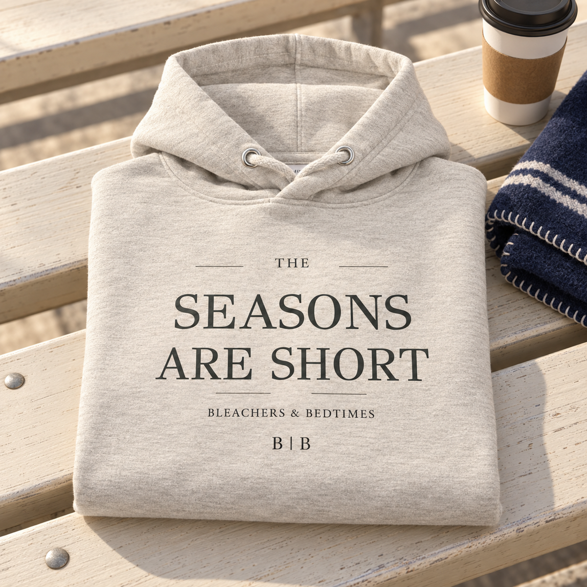 Folded oatmeal “The Seasons Are Short” hoodie on outdoor bleachers beside a coffee cup and navy blanket.