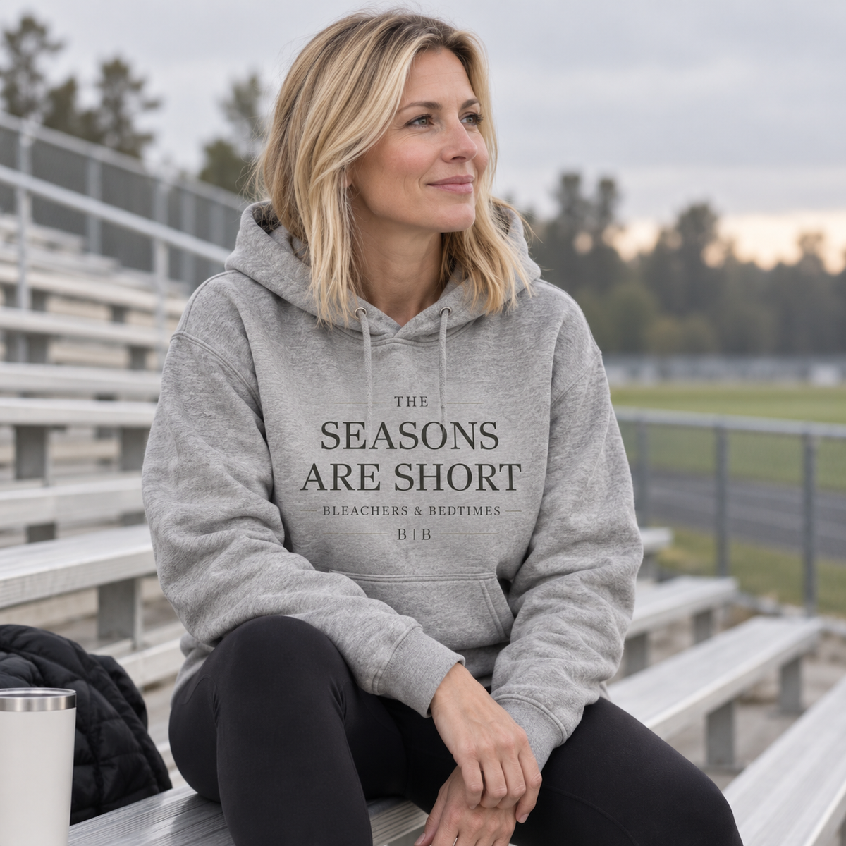 Woman seated on outdoor sports bleachers wearing a carbon gray “The Seasons Are Short” hoodie.