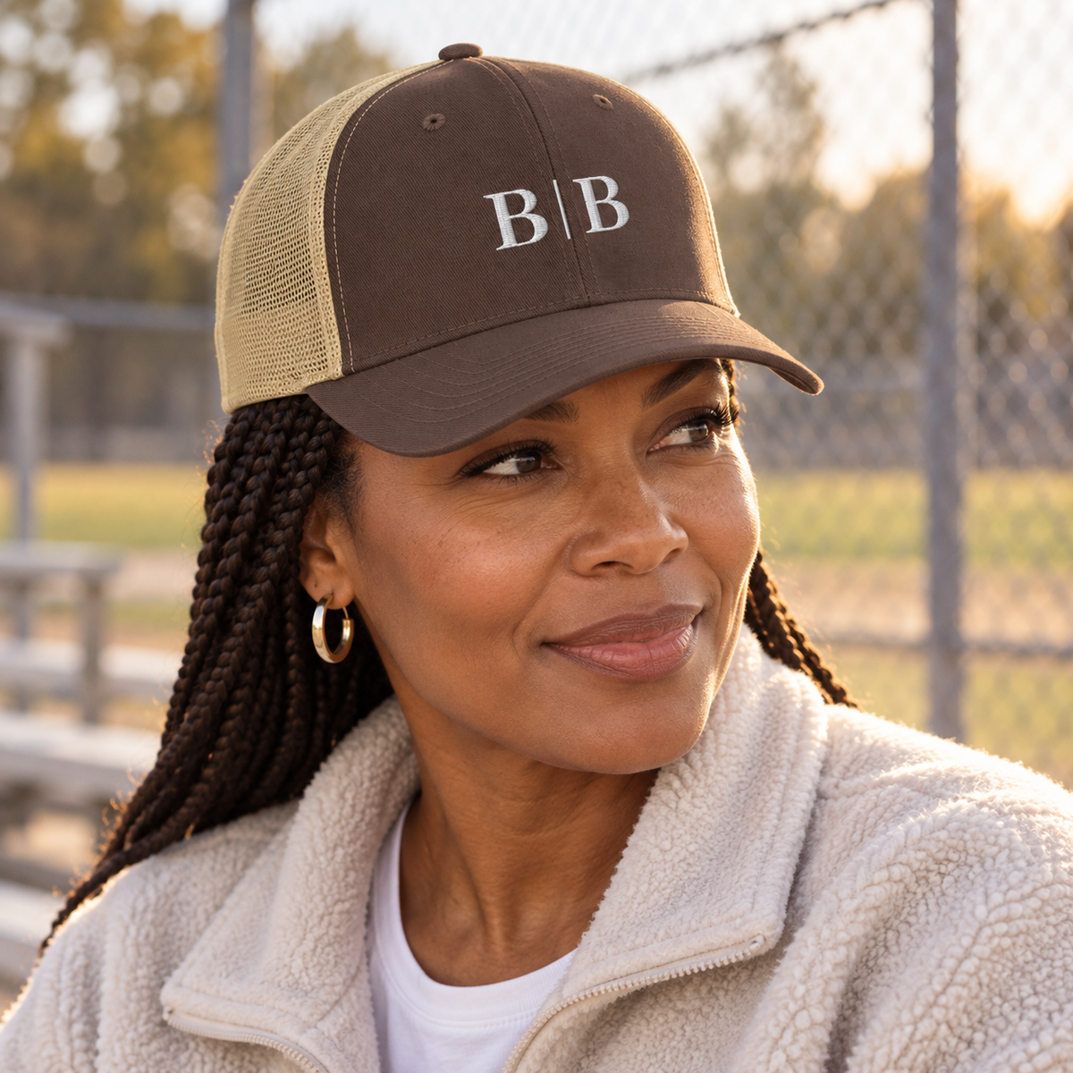 Woman wearing a brown and khaki B | B embroidered trucker hat beside an outdoor sports field.