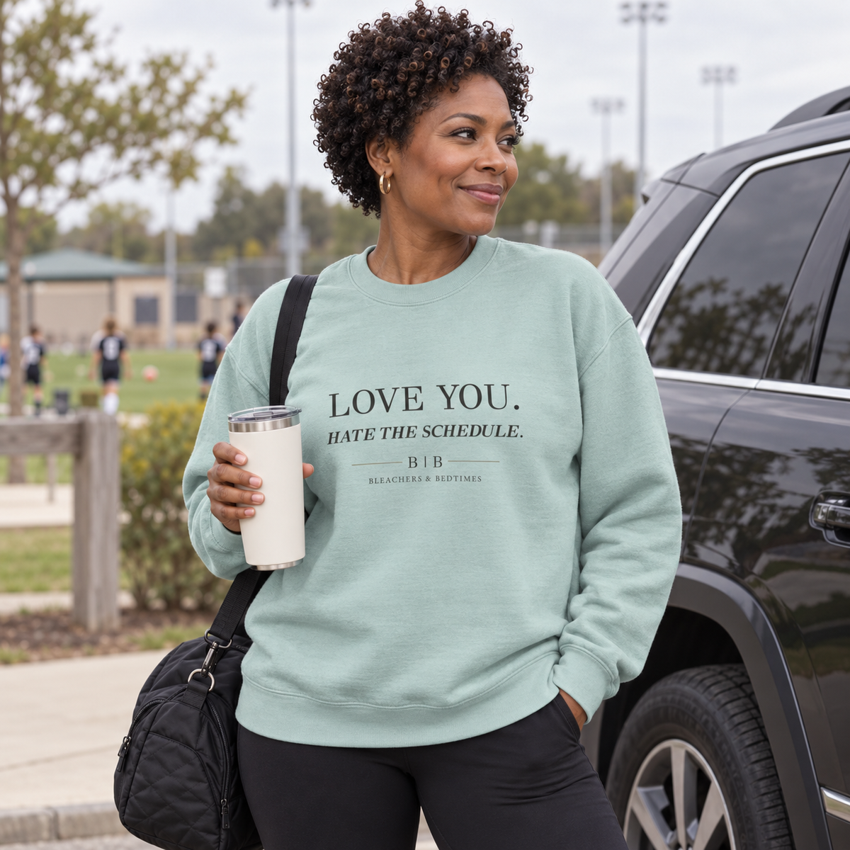 Woman wearing an agave “Love You. Hate the Schedule.” crewneck beside an SUV at a youth sports field.